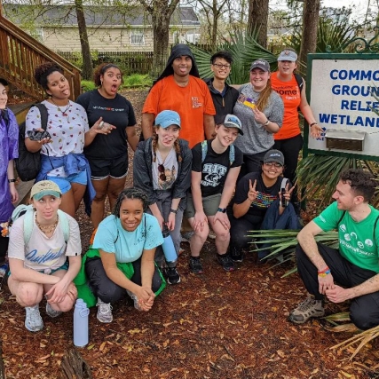 group of 13 students poses outdoors next to sign for Common Ground Relief Wetlands.