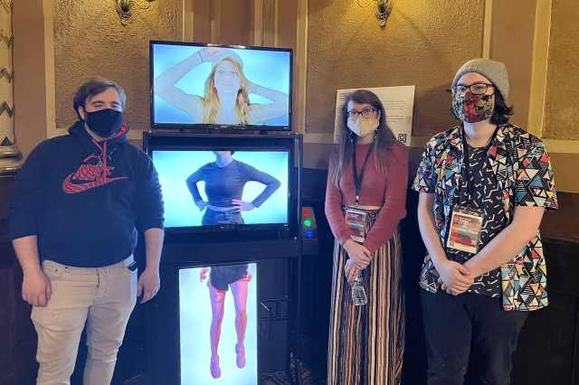 Three people stand around Amalgamate, a three-channel video display.