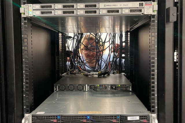 A person wearing glasses behind a server rack peeks through a tangle of cords.