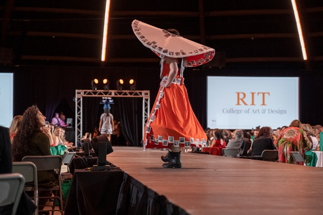 student walking on a runway with a large sombrero style hat and an orange skirt.
