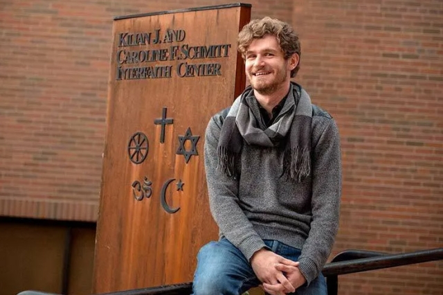 student posing outside next to sign for Kilian J. and Caroline F. Schmitt Interfaith Center.