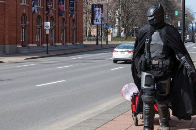 a person dressed as Batman walking down the sidewalk of a city street pulling a wagon.
