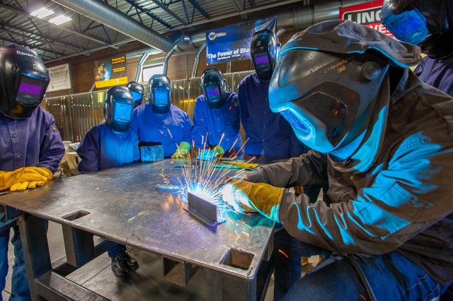 Students gather around table watching welding in blue safety suits.