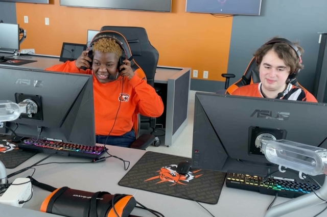 two college students wearing gaming headsets and sitting at a computer station.