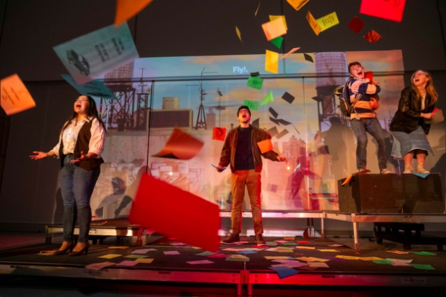 three college students singing on stage with playbills flying through the air. 
