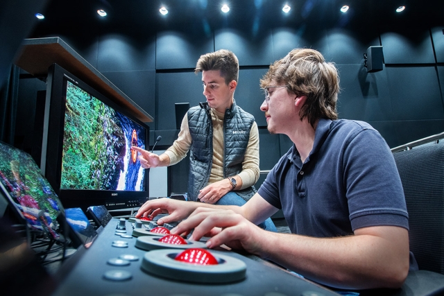 two men using round red joysticks looking at a colorful image on a T V screen.