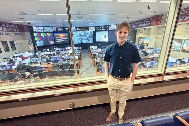 college student standing outside of a glass wall that looks down into a large computer lab.