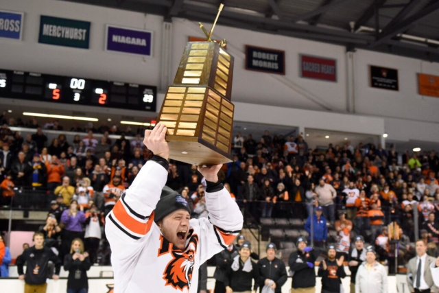an RIT mens hockey player is shown raising the divisional championship trophy above his head as he skates around the gene polisseni center.