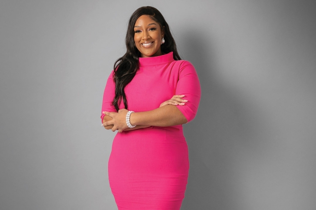 Shakierah Smith is shown standing in front of a gray wall wearing a hot pink dress with her hands crossed across her torso.