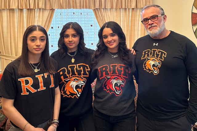 a family stands in a livingroom all wearing R I T shirts.