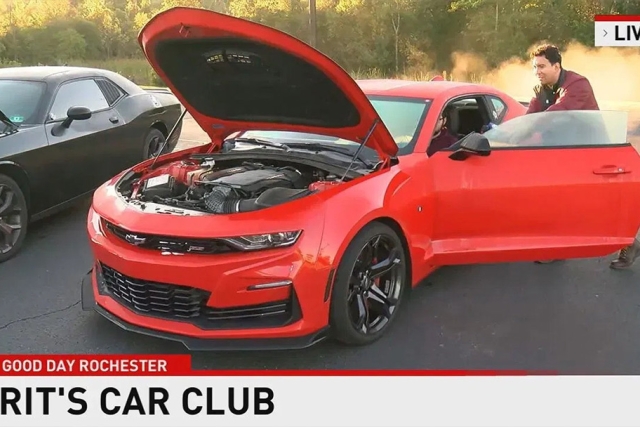 A red dodge charger is the focal point of the image, it's driver side door and hood are open and on view. There is one man sitting in the driver seat and another man standing beside him outside of the car.