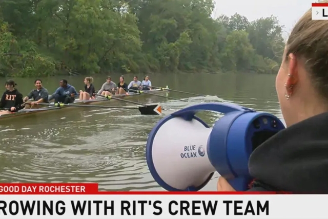 8 members of the RIT rowing team are rowing down the river as part of their practice. There is lush greenery in the background on the other side of the river, and in the foreground of the image there is the side profile of one of the team members using a megaphone to direct her teammates. Along the bottom is a news graphic that reads, Good Day Rochester, Rowing with RIT's Crew Team.
