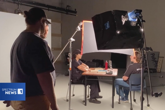 four students sit on a film set. Two of them sit at a table in front of studio lighting, two stand to the side looking at the set.