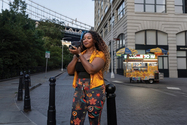Chris Wairegi stands on a street corner in New York City with a smile on her face and hands clasped near her face.