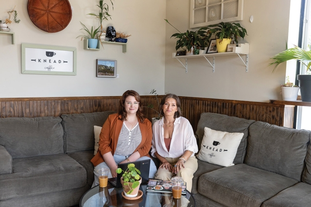 two women sit on a gray couch in a white livingroom with art on the walls.