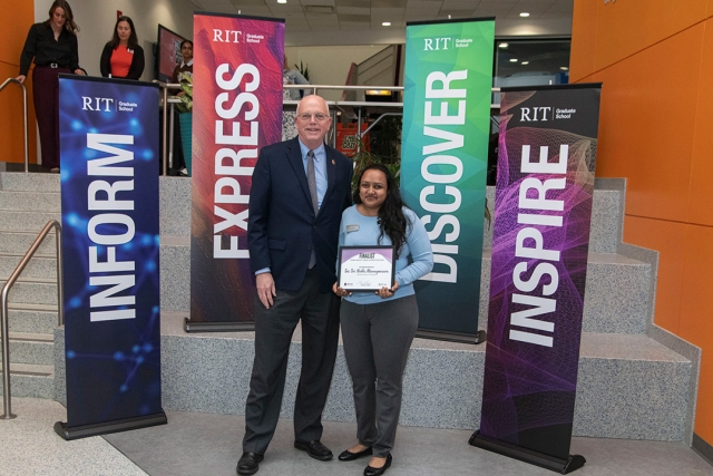 a young indian woman stands beside R I T president David Munson holding a plaque in front of a set of cement steps..