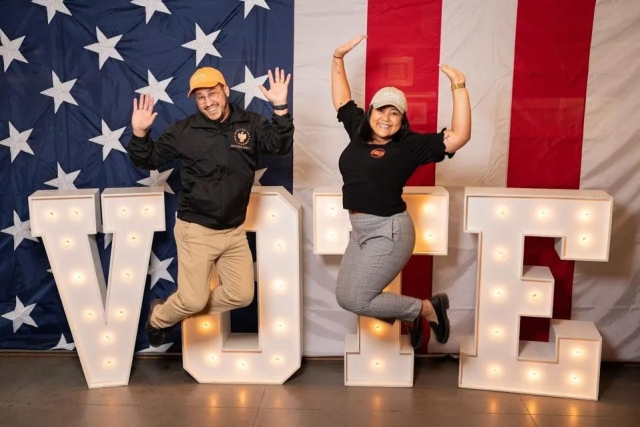 a male and female student jump up in front of a lighted vote sign in front of an american flag.