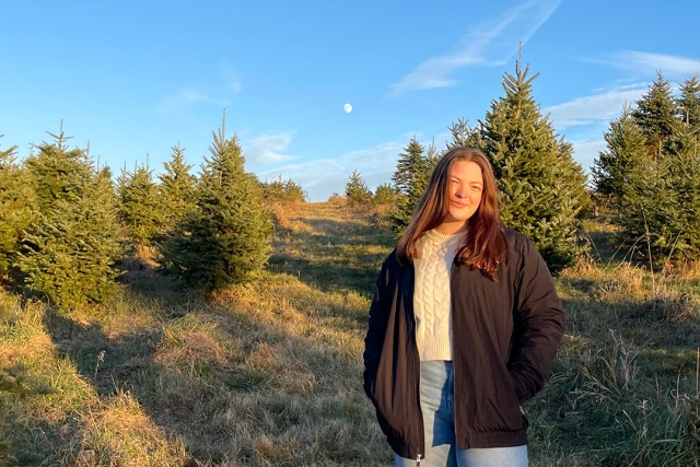 a college age girl standsin a black jacket and cream sweater in a forest area.