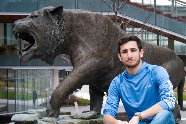 Kyle Scher kneels in front of a tiger statue on campus wearing a long sleeve blue shirt.
