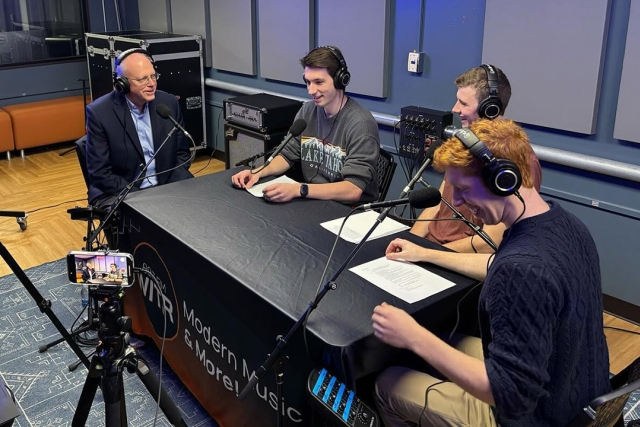 4 people sit around a black table with headphones in front of microphones.