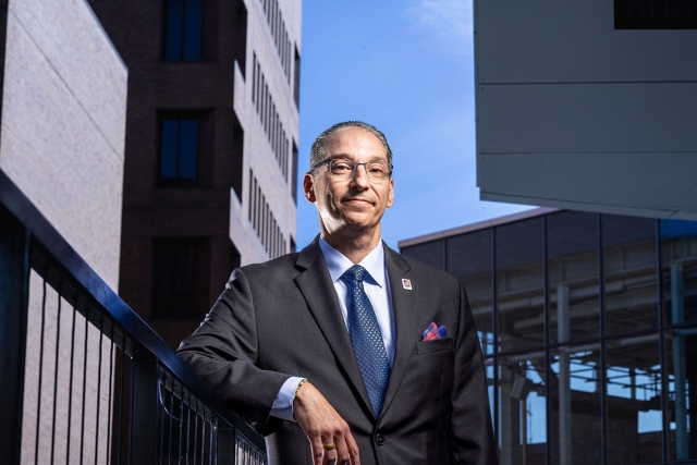 a man leans on a black metal balcony in the midst of brick buildings.