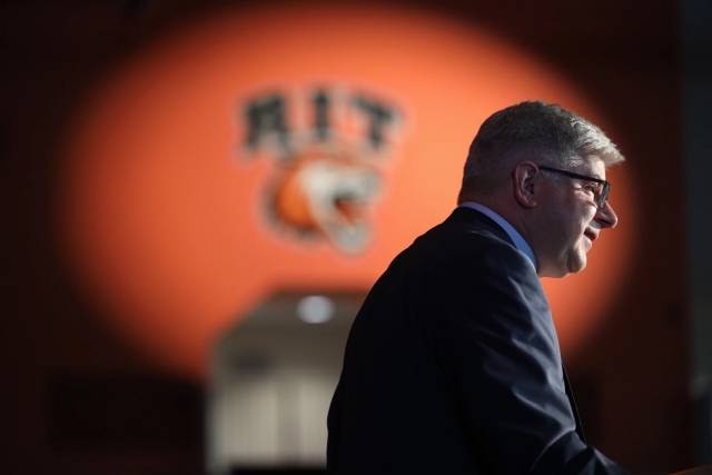 Next RIT president speaks at podium with orange logo lighted in the background