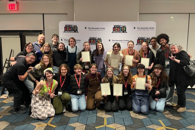 A group of faculty, students and interpreters in two rows, front row kneeling, back row standing, all smiling, several holding framed awards. 