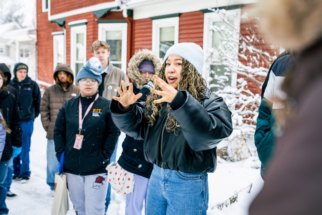 a college age female has her hands up in a circular fashion. She stands in a snowy area with other students and a red house behind her.