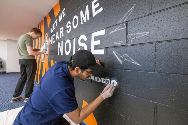 Two artists paint a bold mural on a black brick wall featuring the phrase 'LET ME HEAR SOME NOISE' in large white letters with orange and white graphic elements. One artist in a navy blue shirt and cap details the design, while the other in a green shirt adds finishing touches.