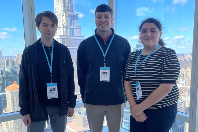 Three students wearing name badges and lanyards pose in front of a large window with a city skyline in the background.