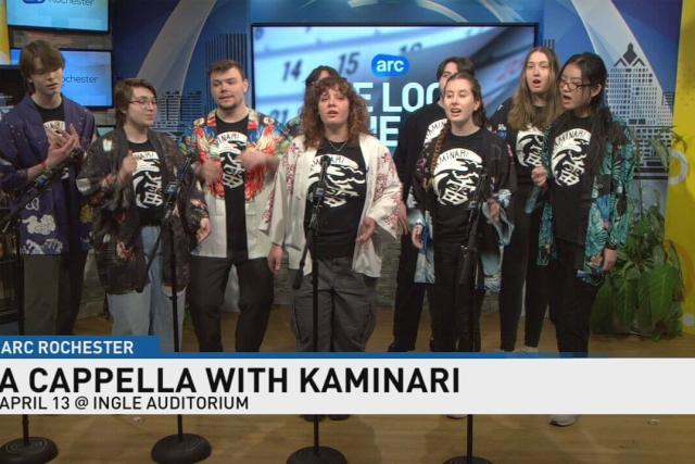 members of an a cappela group stand in front of a news studio background with mics setup in front of them to sing.