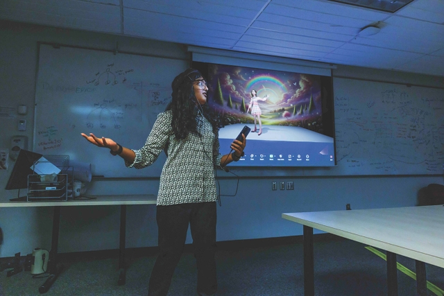 a female stands in a dimly lit classroom holding a phone in one hand. A screen showing a virtual reality game is in the background.