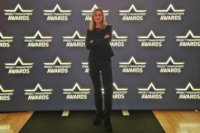 Woman in a black suit stands confidently at the Project Management Awards backdrop as nominee, winner, and attendee.