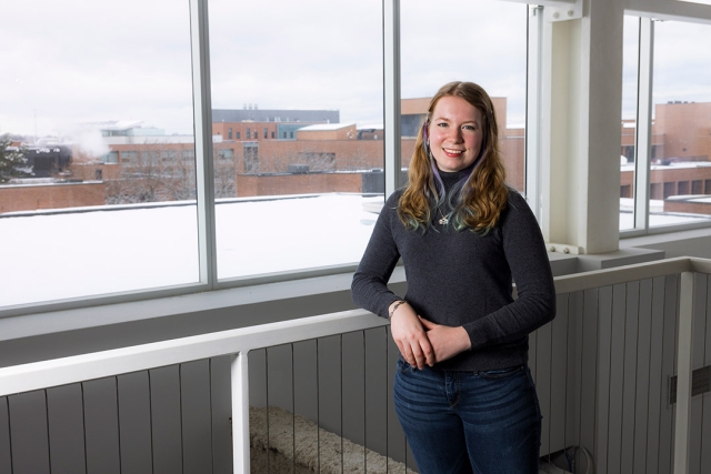 a young woman dressed in jeans and a black sweater looks at the camera and leans on a railing next to windows looking out on to a snow covered campus.