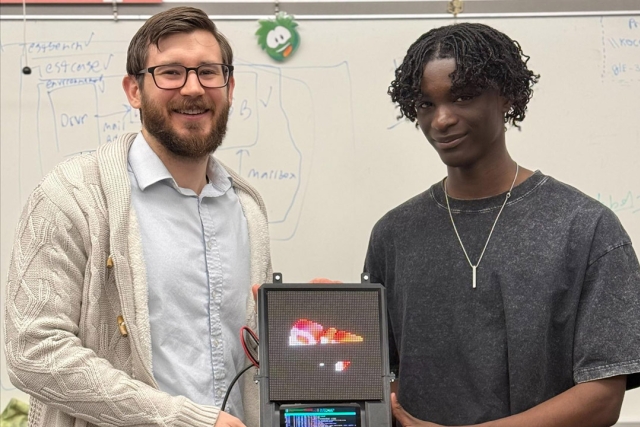 Two people smiling and holding a digital display showing a pixelated fish image in a classroom.