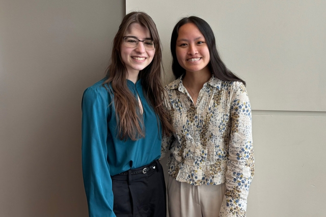 Two college age females stand in front of a beige colored wall. The girl on the left has long brown hair and wears a blue shirt with black pants and the other has long black hair and wears a floral top with beige pants.