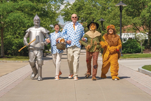 Dave Munson and Nancy Munson, dressed in blue gingham suits, walk down a sunny pathway with other adults dressed in Wizard of Oz costumes.