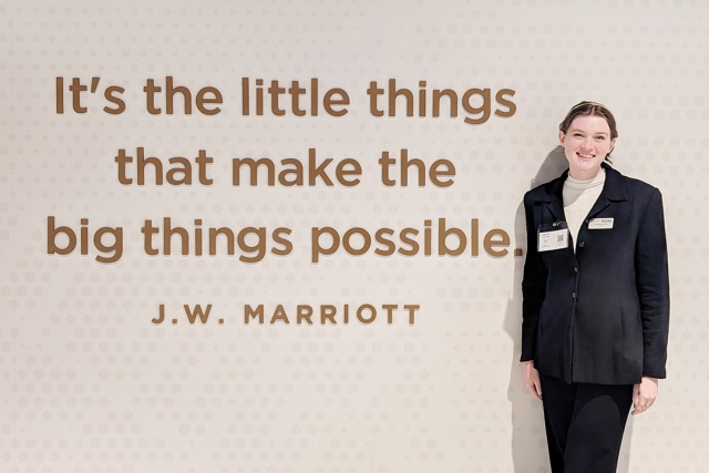 a college age woman in a black suit stands next to a wall displaying a quote by J W Marriott that says It's the little things that make the big things possible.