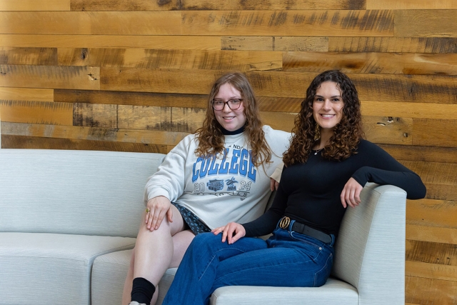 Two women sit smiling on a white couch in front of a wooden wall.