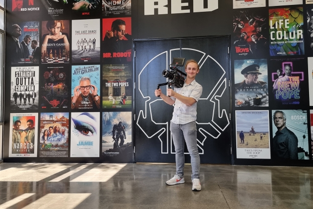 Max Schaefer holds a camera, with many movie posters behind him.