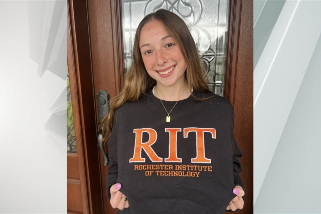 Young woman stands in front of house door showing off her RIT Rochester Institute of Technology shirt.