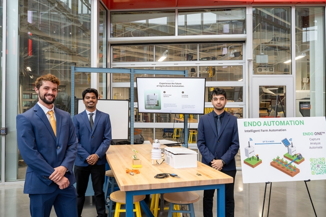 Three men in suits stand by agricultural automation display table and presentation boards.
