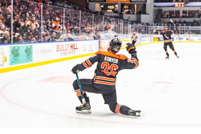 A hockey player takes a knee on the ice and faces the crowd, giving the cheering group a wave. There is another RIT hockey player in the background.