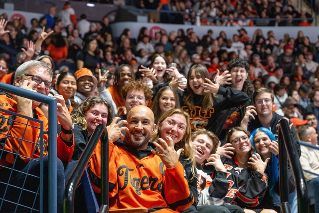 Fans cheer on the men’s hockey team at the Blue Cross Arena. A large group of people in RIT branded clothes smile at the camera doing the ASL sign for tiger.