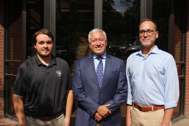 three men stand in front of a building smiling at the camera.