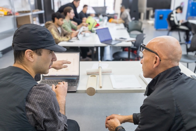 a student in a black hat speaks with a man sitting to his right, gesturing near a laptop screen.