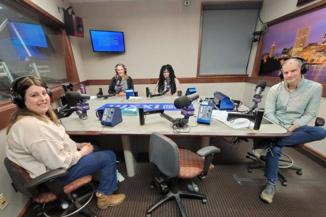 Four people wearing headphones sit at a table in a radio talk studio: a woman front left has long brown hair and is wearing a beige shirt, blue jeans and brown boots; a man front right has short dark hair and is wearing a light green button-down shirt, blue jeans and sneakers; a woman back left has long blonde hair and is wearing a black zip-up fleece over a grey t-shirt; a woman back right has long black and green dreadlocks and is wearing glasses and a pink shirt.
