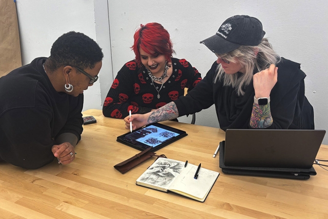 Three students collaborate around a tablet at a classroom table, reviewing digital artwork alongside sketchbooks and a laptop.