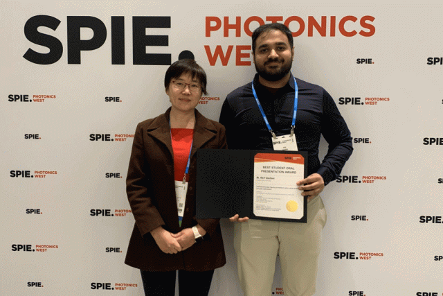 Two people stand in front of a banner, with man on right holding a certificate.