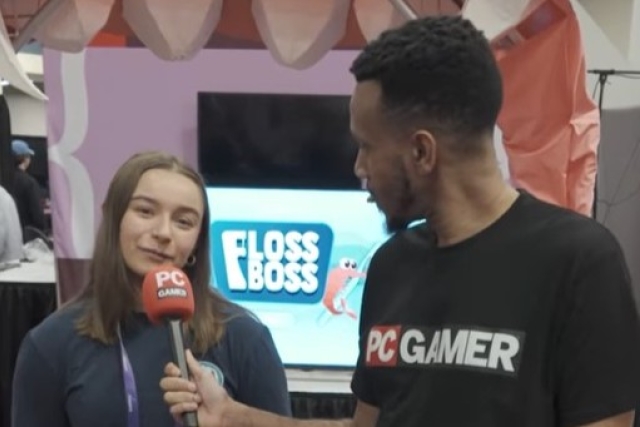 a girl stands in front of a TV screen displaying the words Floss Boss as she is interviewed by a man in the P C Gamer shirt.